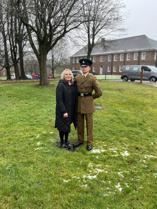 A person in a black coat stands beside a person in a military uniform. They are outside on a grassy area with a brick building, leafless trees, and parked vehicles in the background on a cloudy day.