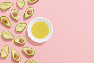 Halved avocados on the left of a pink background. A white bowl filled with yellow oil